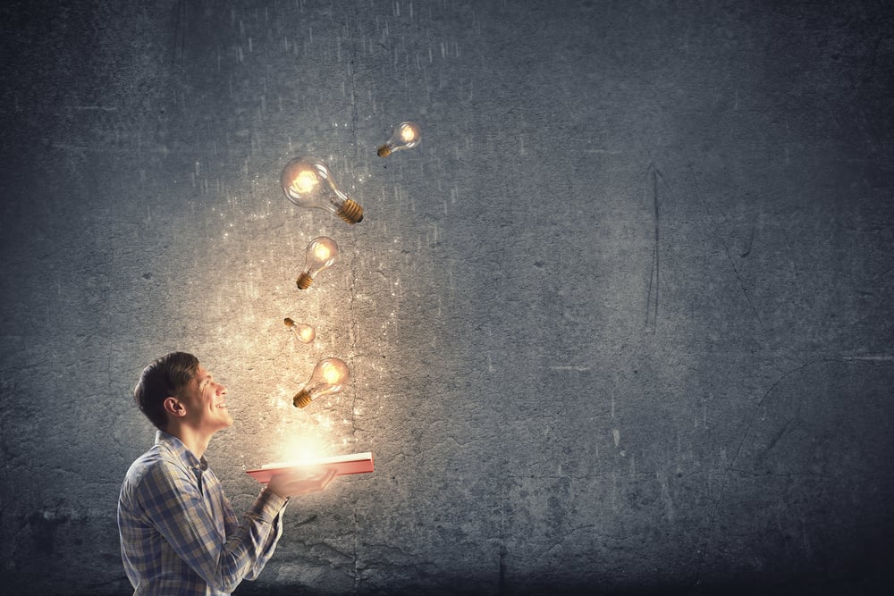 Young man holding opened book with glass glowing light bulbs flying out